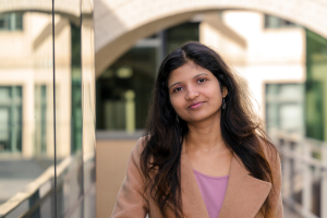 Keerthi Kunnumkai leans against a railing outside of a glass building and smiles at the camera.