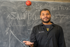 Byron Daniel stands in front of a blackboard with physics equations written on it. He throws an apple in the air as he smiles at the camera.