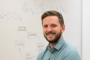 Ben Koby stands in front of an out-of-focus whiteboard with chemistry formulas on it and smiles at the camera.