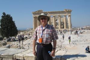 Victor Bearg stands in front of the Acropolis in Greece and smiles at the camera. He's wearing a plaid shirt, a hat and sunglasses.