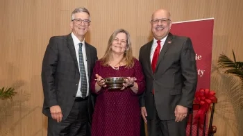 A woman stands between two men holding a broad silver cup with two handles.