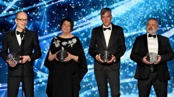 Three men and one woman hold spherical wire sculptures on a stage. The background is blue and filled with flashes of light.