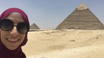Smiling woman wearing sunglasses and a purple headscarf, with a desert landscape and two pyramids visible in the background.