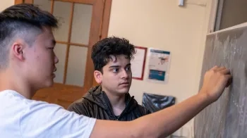 Two male students in a classroom setting; one is writing on a blackboard while the other observes nearby, both dressed casually and engaged in the activity.