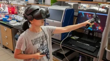 A woman wearing a virtual reality headset moves her arms while a monitor shows the interior of an infected cell that she is navigating.