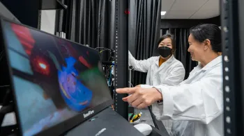 Kate Hong shows an image on a screen to a graduate student. Both are wearing lab coats. Kate Hong is smiling. The student wears a face mask.